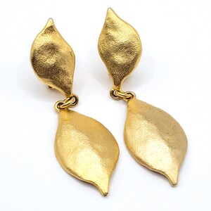 May include: A pair of gold-coloured leaf-shaped earrings. Each earring features a textured, elongated top piece connected to a larger, smooth leaf-shaped dangle. The earrings have a clip-on style.