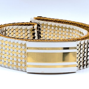 May include: A white and gold metal belt with a white buckle featuring two gold stripes. The belt is made of interlocking metal scales.
