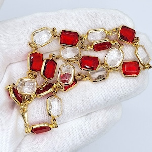 May include: A gold necklace with alternating red and clear rectangular gemstones. The gemstones are set in a gold frame and linked by delicate gold chains. The necklace is displayed on a white surface.