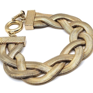 May include: A gold-toned bracelet with a braided design. The bracelet features a series of interwoven, textured bands, creating a thick, statement piece. It has a rectangular clasp and a circular toggle closure. The jewelry is set against a white background.