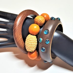 Puede incluir: Tres brazaletes de madera con un diseño de cuentas. Los brazaletes son marrones y tienen un acabado de madera natural. Las cuentas son de color naranja, amarillo y marrón. Los brazaletes están apilados uno encima del otro.