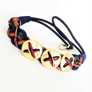 May include: A braided navy blue, red, green, and orange rope belt with three gold-toned buttons. The buttons have a cross-shaped design.