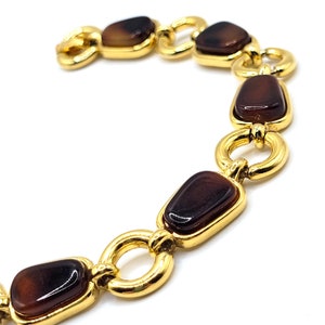 May include: A gold-toned chain bracelet featuring rectangular, dark brown, glossy stones. The stones are set within a gold frame and connected by gold ring-shaped links. The bracelet is displayed against a white background.
