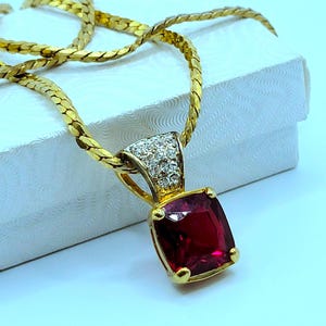 May include: A gold-toned necklace featuring a thick, flat herringbone chain and a pendant with a large, square-cut deep red gemstone. The pendant bail is accented with small, clear, sparkling stones set in a gold-toned metal frame.