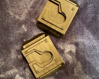 May include: Two small, rectangular, metallic, gold-colored objects with a raised, geometric design. The objects have a textured surface and a small, circular hole in the center.