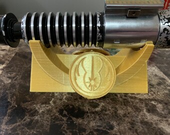 May include: A gold-colored plastic stand with a circular cutout featuring a stylized lightsaber design. The stand is designed to hold a lightsaber, which is visible in the background.