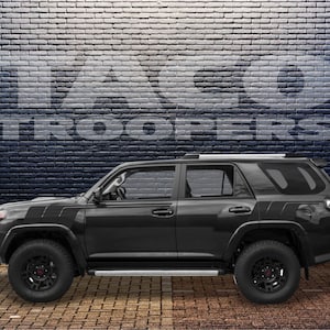 Puede incluir: Un SUV Toyota 4Runner negro con ruedas negras y un portaequipajes de techo negro. El vehículo está estacionado frente a una pared de ladrillo con las palabras "Taco Troopers" escritas en ella.