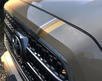 Könnte beinhalten: Nahaufnahme eines beigen Lkw mit einem schwarzen Kühlergrill und einem verchromten Toyota-Emblem. Der Lkw hat einen dreistufigen Rennstreifen auf der Motorhaube in Braun, Gold und Silber.