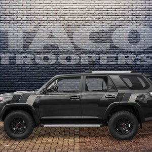 May include: A black Toyota 4Runner with grey and black stripes on the sides. The vehicle is parked in front of a brick wall with the words "Taco Troopers" written on it.