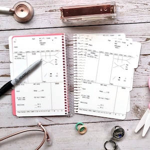 May include: A white medical record keeping binder with pink edges and a spiral binding. The binder has a section for patient information, vital signs, and medications. A black pen is lying on top of the binder.