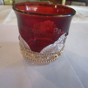 May include: A ruby red glass with the etched text "Thompson Pa." is held by a decorative, clear glass holder with gold trim. The glass has a rounded shape and a gold rim.