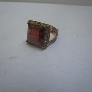 May include: A gold ring with a large, square, red gemstone surrounded by smaller clear gemstones.