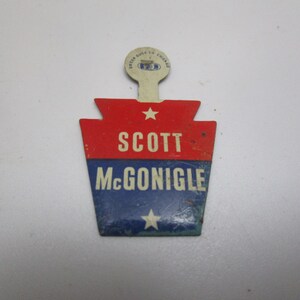 May include: A red, white, and blue campaign button shaped like the state of Pennsylvania. The button features the text "SCOTT McGONIGLE" and two white stars.