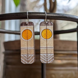 Op de afbeelding: Een paar houten oorbellen met een geometrisch ontwerp. De oorbellen zijn geschilderd in tinten bruin, wit en geel. De oorbellen hebben een cirkel met een gele kern en een witte omtrek.