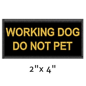 May include: A rectangular black patch with gold embroidered text that reads "WORKING DOG DO NOT PET." The patch measures 2" x 4" and has a decorative border. This patch is designed to be attached to a working dog's gear.