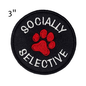 May include: A 3-inch diameter black patch with white text that reads "SOCIALLY SELECTIVE". A red paw print is in the center. The patch is circular and has a woven texture.