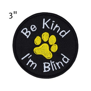 May include: A round, black embroidered patch with the text "Be Kind I'm Blind" in white. A yellow paw print is centered below the words "Be Kind". The patch is 3 inches in diameter.