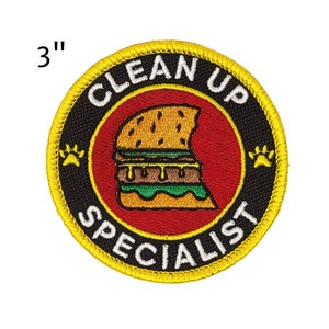May include: Embroidered patch, 3 inches in diameter, with a yellow border and the words "CLEAN UP SPECIALIST" in white. The patch features a red center with a cartoon burger and paw prints.