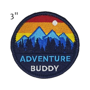 May include: A 3-inch round embroidered patch with a mountain and sunset design. The patch features a blue mountain range, a yellow and orange sunset, and the text "ADVENTURE BUDDY" in white.