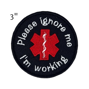 May include: A 3-inch round patch with the text "Please ignore me I'm working" in white lettering. The patch has a black background with a red medical symbol in the center.