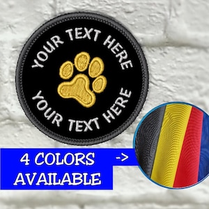 May include: A black, round embroidered patch with a gold paw print in the center. The text "YOUR TEXT HERE" is embroidered in white around the paw print. A small inset shows four color options. The text "4 COLORS AVAILABLE" is in a blue rectangle.