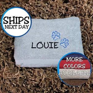 May include: A light blue towel with the name "LOUIE" embroidered in black, along with two blue paw prints. The towel is displayed on a bed of brown packing material. The text "MORE COLORS AVAILABLE" is visible.