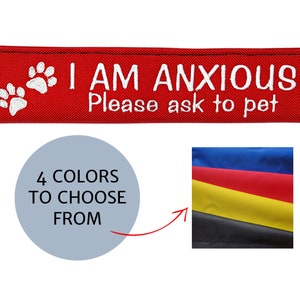 May include: Red dog collar attachment with white embroidered text: "I AM ANXIOUS Please ask to pet." Features paw prints. Includes a graphic with "4 COLOURS TO CHOOSE FROM" and a colour swatch of blue, red, yellow, and black.