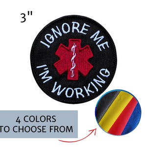 May include: A 3-inch round black patch with white embroidered text that reads "IGNORE ME I'M WORKING". A red medical symbol is in the center. The image also shows a color palette with four color options.