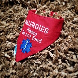 May include: A red dog bandana with white embroidered text that reads "ALLERGIES Please Do not feed." A blue medical symbol is embroidered below the text. The bandana is triangular and rests on a bed of brown paper.