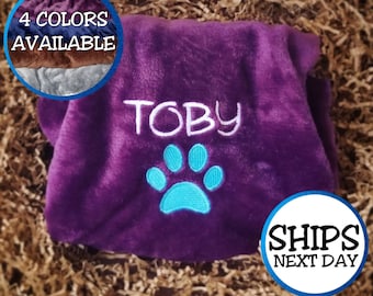 May include: A plush, purple pet blanket embroidered with the name "TOBY" in white and a turquoise paw print. The blanket is surrounded by a circular image showing 4 color options. The text "4 COLORS AVAILABLE" is also visible.