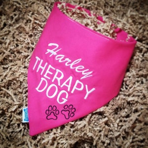 May include: A bright pink dog bandana with white embroidered text that reads "Harley THERAPY DOG". Two black paw prints are below the text. The bandana is made of fabric and has a tag that says "Furgoous".