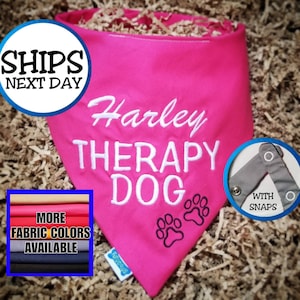 May include: A bright pink dog bandana embroidered with "Harley THERAPY DOG" in white script. Features paw prints and a snap closure. Additional fabric colors are available.