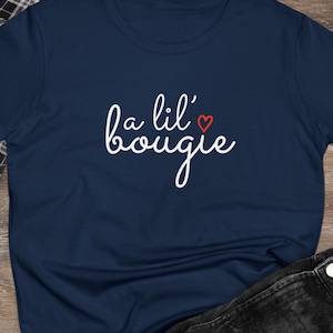 May include: A navy blue short-sleeved t-shirt featuring the white cursive text "a lil' bougie" with a small red heart icon above the letter i. The shirt is displayed alongside a black and white plaid flannel shirt, black denim jeans, and sneakers.