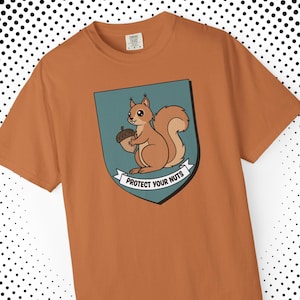 May include: A rust-colored t-shirt featuring a cartoon squirrel holding an acorn within a teal shield. The shield has a banner that reads "PROTECT YOUR NUTS". The squirrel is brown with a fluffy tail.