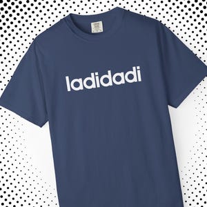 May include: A navy blue t-shirt featuring the white text "ladidadi" across the chest. The top has a crew neck and short sleeves. The background is a white and black polka dot pattern.