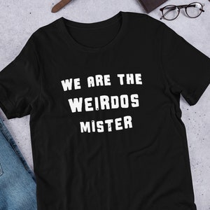 May include: A black short-sleeved t-shirt featuring the white distressed-style text "WE ARE THE WEIRDOS MISTER" printed in the center of the chest. The garment has a crew neckline and a standard fit, showcasing a simple and clean design.