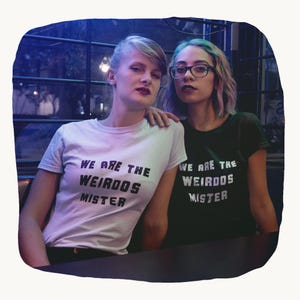 May include: A light-colored t-shirt and a dark green t-shirt, both featuring the black text "WE ARE THE WEIRDOS MISTER" printed on the front. The shirts have short sleeves and a crew neckline, displaying a consistent graphic design across both garments.