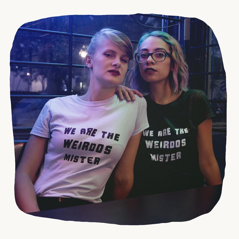 May include: A light-colored t-shirt and a dark green t-shirt, both featuring the black text "WE ARE THE WEIRDOS MISTER" printed on the front. The shirts have short sleeves and a crew neckline, displaying a consistent graphic design across both garments.