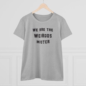 May include: A heather gray short-sleeve t-shirt with a crew neckline. The front of the shirt features the black text "WE ARE THE WEIRDOS MISTER" printed in a bold, distressed, sans-serif font. The garment is displayed on a wooden hanger.