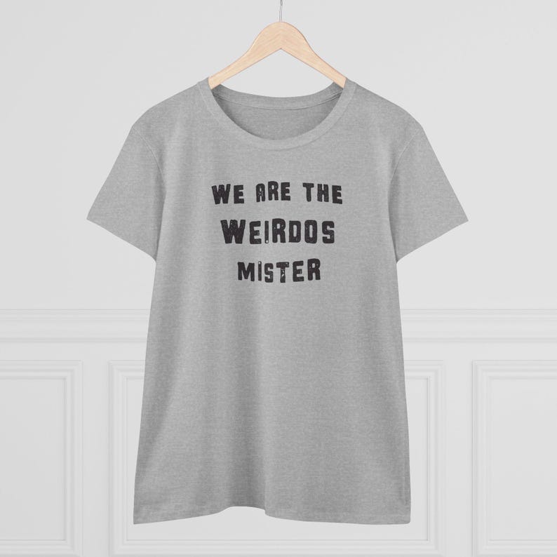 May include: A heather gray short-sleeve t-shirt with a crew neckline. The front of the shirt features the black text "WE ARE THE WEIRDOS MISTER" printed in a bold, distressed, sans-serif font. The garment is displayed on a wooden hanger.