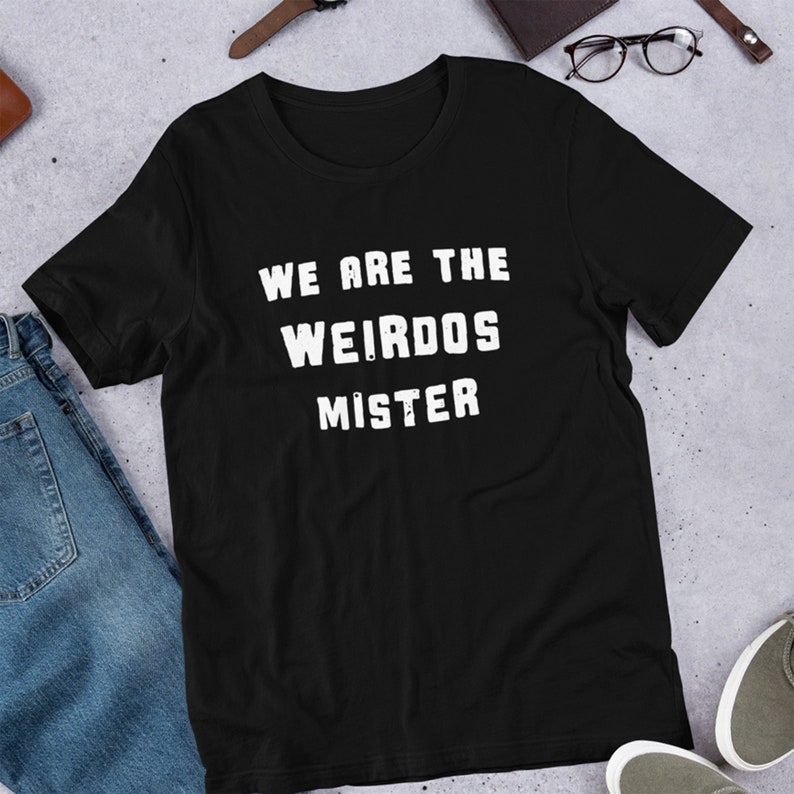 May include: A black short-sleeved t-shirt featuring the white distressed-style text "WE ARE THE WEIRDOS MISTER" printed in the center of the chest. The garment has a crew neckline and a standard fit, showcasing a simple and clean design.