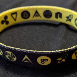 May include: Black and yellow woven wristband with various cryptocurrency logos printed on it.