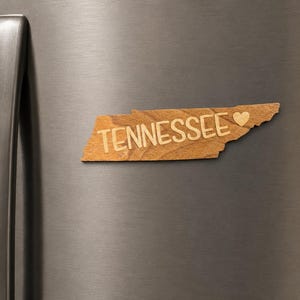 May include: A wooden magnet shaped like the state of Tennessee with the word TENNESSEE and a small heart symbol engraved on the front. The magnet has a natural light brown wood grain finish and is attached to a metallic gray refrigerator door.
