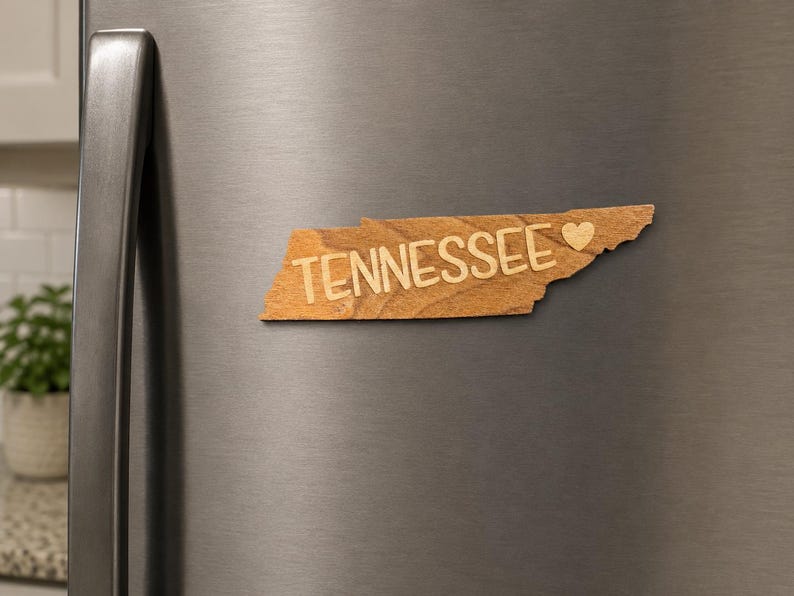 May include: A wooden magnet shaped like the state of Tennessee with the word TENNESSEE and a small heart symbol engraved on the front. The magnet has a natural light brown wood grain finish and is attached to a metallic gray refrigerator door.