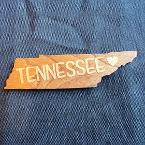 May include: A wooden cutout shaped like the state of Tennessee with the word TENNESSEE printed in a light-colored sans-serif font across the center, accompanied by a small heart symbol positioned near the eastern border of the state shape.
