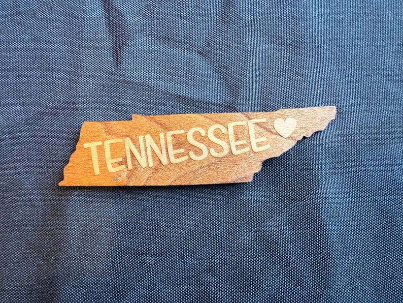 May include: A wooden cutout shaped like the state of Tennessee with the word TENNESSEE printed in a light-colored sans-serif font across the center, accompanied by a small heart symbol positioned near the eastern border of the state shape.