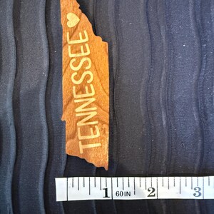 May include: A light brown wooden cutout shaped like the state of Tennessee with the word TENNESSEE and a small heart symbol engraved on the surface. A white measuring tape is positioned below the object, showing a length of approximately 3 inches.