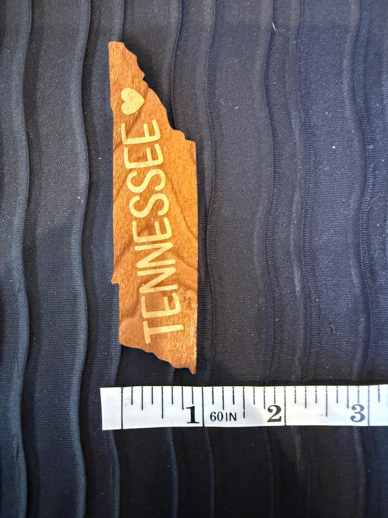 May include: A light brown wooden cutout shaped like the state of Tennessee with the word TENNESSEE and a small heart symbol engraved on the surface. A white measuring tape is positioned below the object, showing a length of approximately 3 inches.