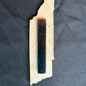 May include: A light-colored wooden cutout shaped like the state of Tennessee with a long, rectangular black magnetic strip attached to the center of the back. The wood features a natural grain pattern and a smooth, flat surface with irregular edges.