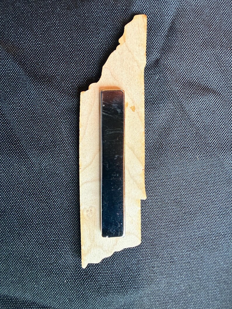 May include: A light-colored wooden cutout shaped like the state of Tennessee with a long, rectangular black magnetic strip attached to the center of the back. The wood features a natural grain pattern and a smooth, flat surface with irregular edges.
